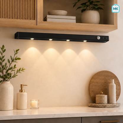 A sleek black under-cabinet LED light bar mounted on a beige wall above a kitchen counter, casting a warm glow downward. The scene includes wooden shelves, plants, and decorative items in a modern kitchen setting.