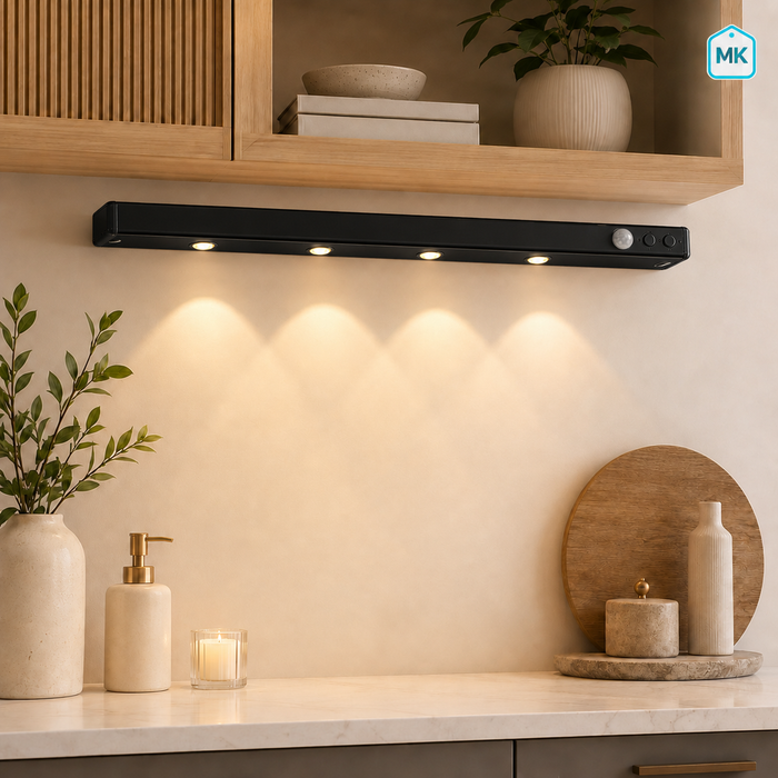 A sleek black under-cabinet LED light bar mounted on a beige wall above a kitchen counter, casting a warm glow downward. The scene includes wooden shelves, plants, and decorative items in a modern kitchen setting.